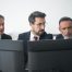 trio of colleagues in dark suits looking at dual-screen workstation